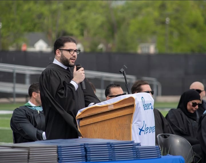 Nasri Sobh speaking from a podium in academic robes at an outdoor high school graduation.
